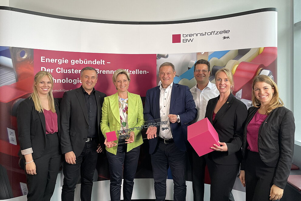 Das Team des Clusters Brennstoffzelle BW posiert gemeinsam mit Baden-Württembergs Wirtschaftsministerin Dr. Nicole Hoffmeister-Kraut. Sie stehen dabei vor einem Informations-Aufsteller des Clusters.