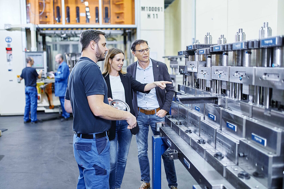 Drei Personen stehen in einer Produktionshalle, einer der Männer hält ein Produktionsteil in der Hand, die Frau zeigt auf die Maschine. 