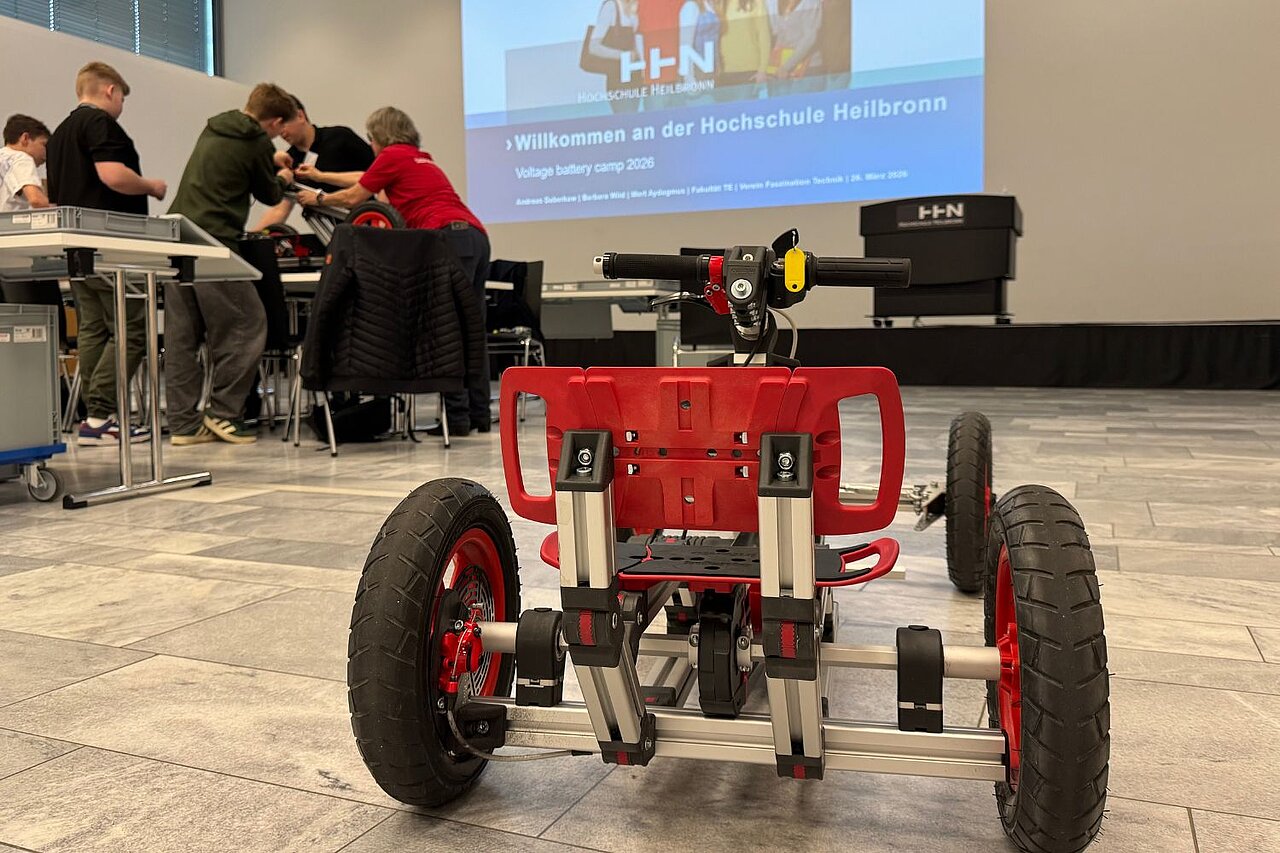 Nahaufnahme und Rückansicht eines Elektro-Karts; im Hintergrund baut eine Gruppe Jugendlicher ein Elektro-Kart zusammen.