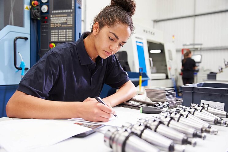 Eine Ingenieurin schreibt, im Hintergrund ist eine CNC-Maschine zu sehen.