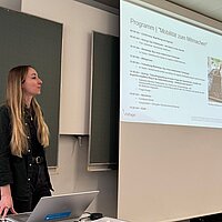 Eine Frau steht vor einer Leinwand und stellt das Programm des Battery Camps vor.