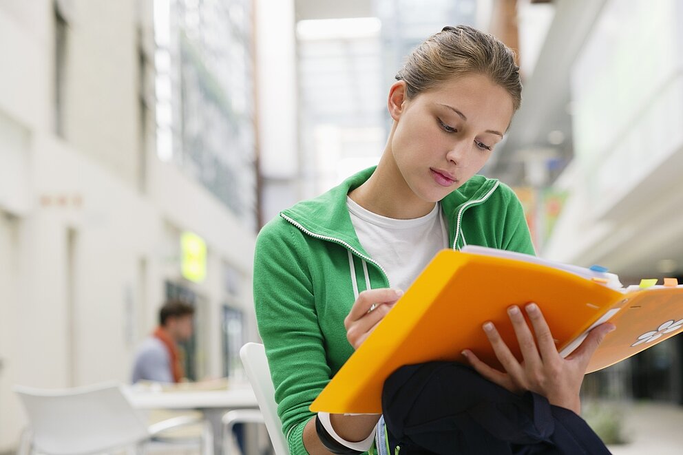Eine junge Schülerin in einem grünen Hoodie ließt in einer orangefarbenen Mappe