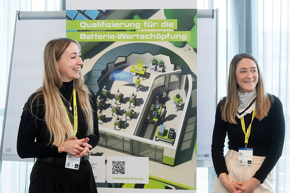 Zwei lächelnde Frauen stehen vor einem Roll-up mit der Aufschrift 'Qualifizierung für die Batterie-Wertschöpfung' und einer schematischen Darstellung eines Batterie-Ökosystems.