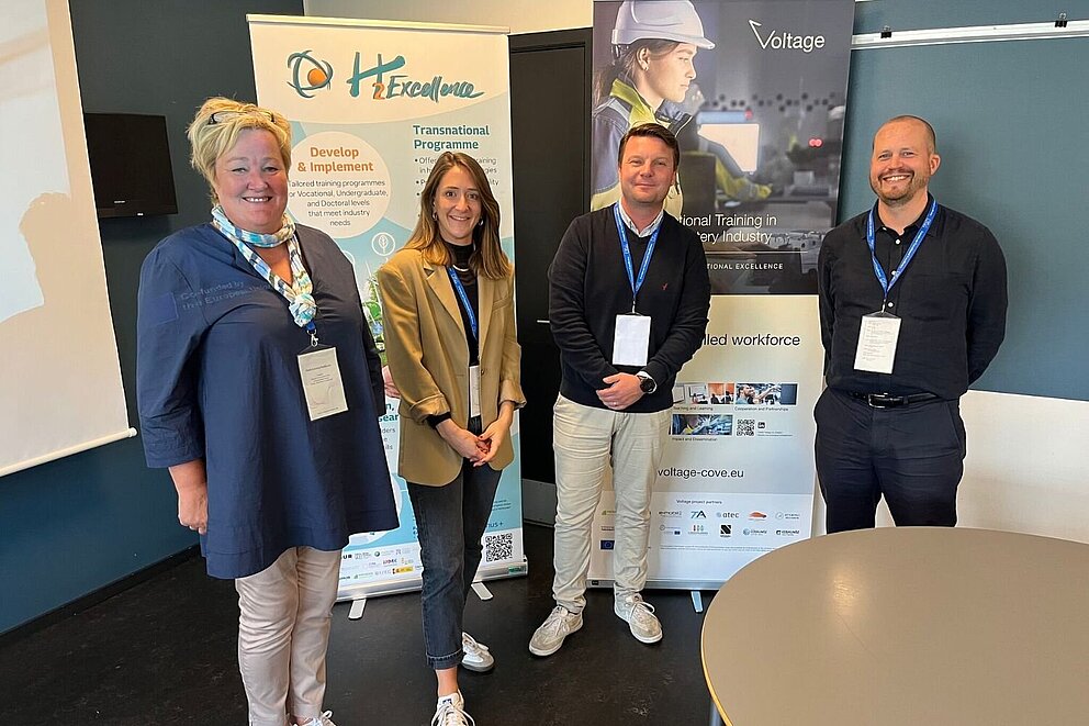 Vier Personen stehen in einem Raum während des Workshops im Rahmen des Forum on Vocational Excellence in Kolding, Dänemark. Links ein Banner des Projekts H2Excellence und rechts ein Banner des Projekts Voltage.