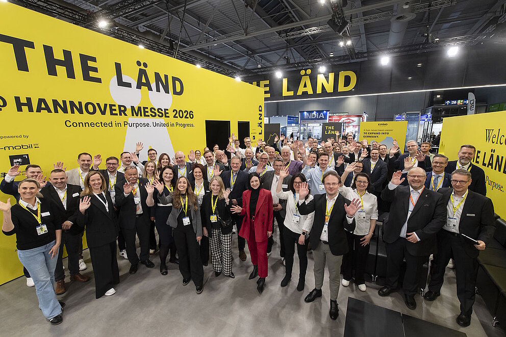 Aussteller des THE LÄND Gemeinschaftsstandes auf der Hannover Messe stehen vor einer gelben Messewand und winken in die Kamera