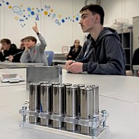 Nahaufnahme runder Natrium-Ionen-Batterien; im Hintergrund sitzen Jungen und hören einem Vortrag zu.