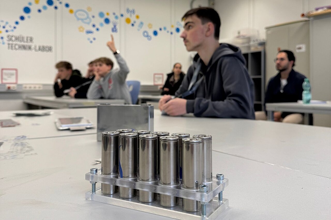Nahaufnahme runder Natrium-Ionen-Batterien; im Hintergrund sitzen Jungen und hören einem Vortrag zu.