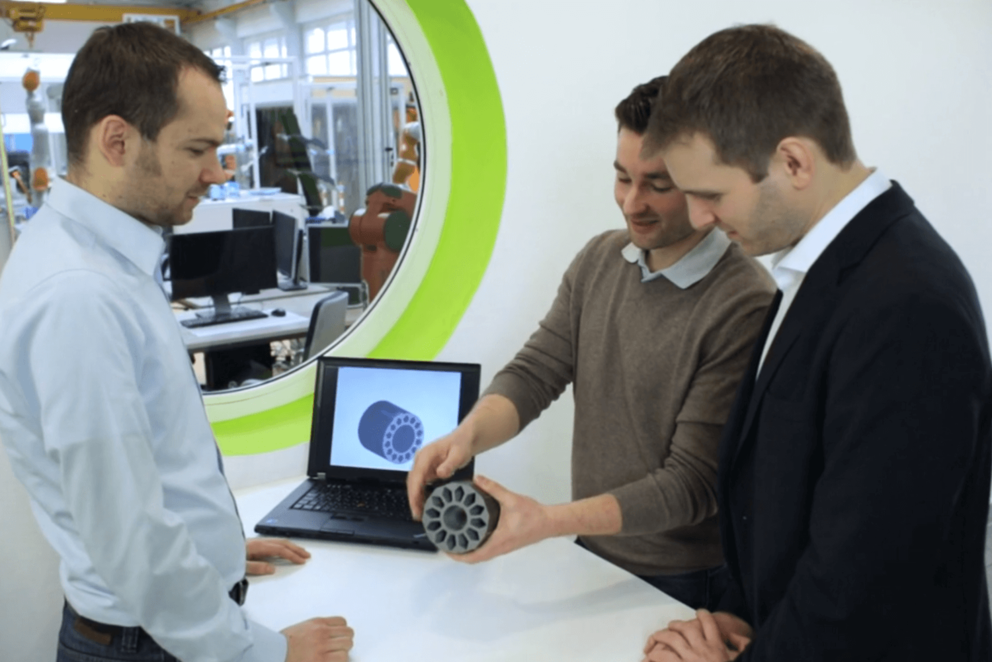 Drei Personen begutachten einen Stator. Im Hintergrund ist eine Produktionshalle zu sehen.
