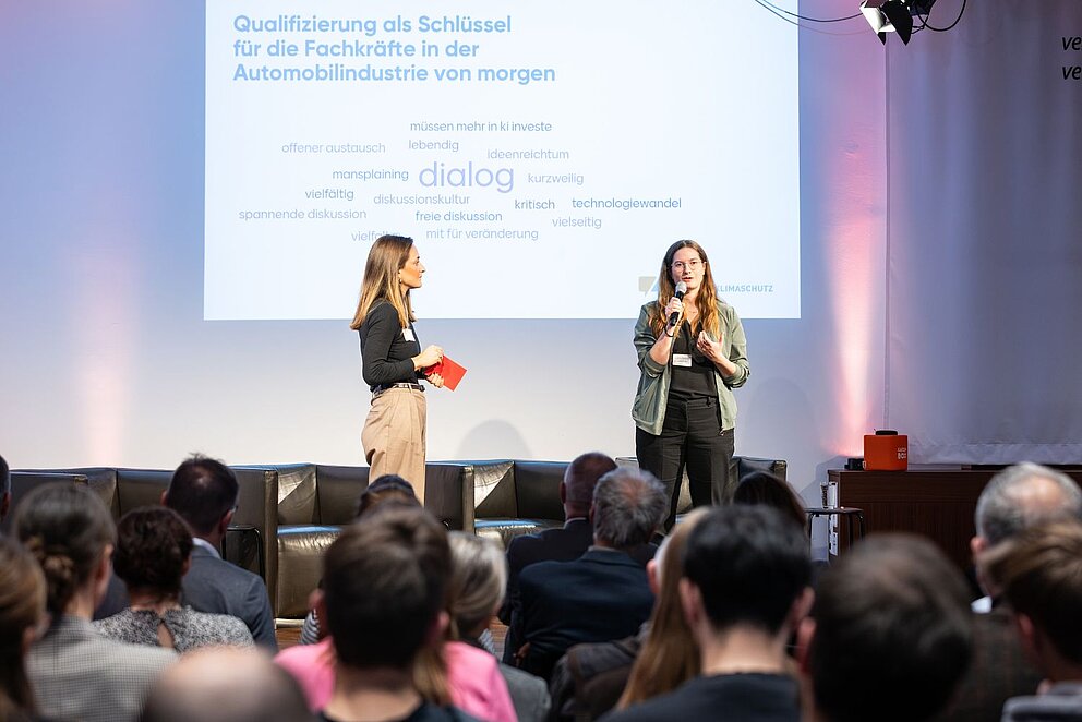 Zwei Frauen stehen auf einer Bühne vor Publikum und halten einen Vortrag mit dem Titel ‚Qualifizierung als Schlüssel für die Fachkräfte in der Automobilindustrie von morgen‘.