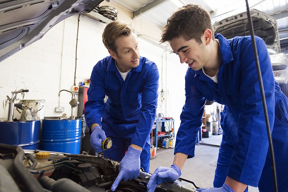 Zwei junge Automechaniker beugen sich über den offenen Motorraum eines Autos in einer Kfz-Werkstatt.