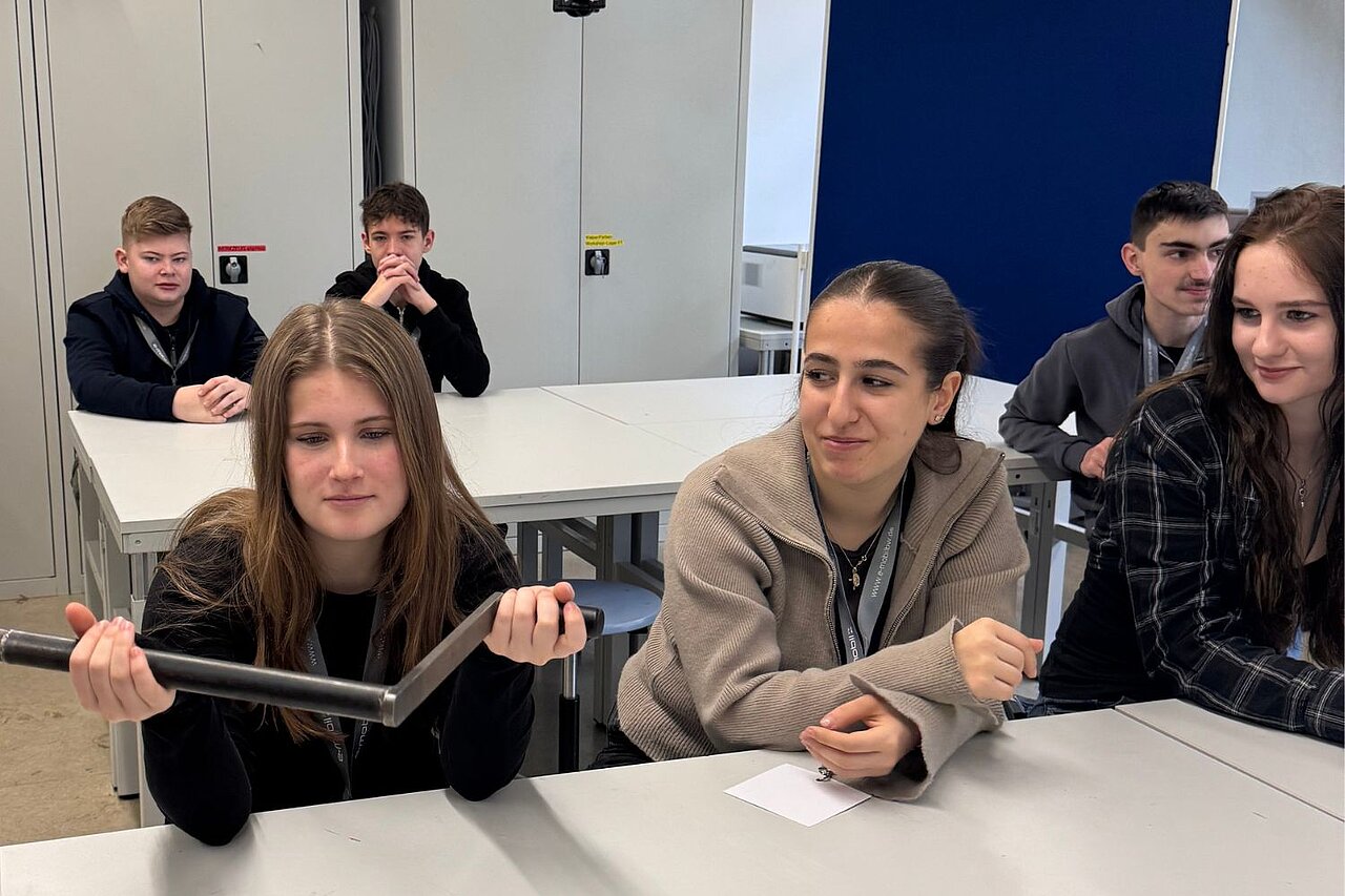 Eine Gruppe Schülerinnen und Schüler sitzt in einem Raum; ein Mädchen hält einen Kurbelanzünden für Autos in der Hand. 