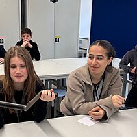 Eine Gruppe Schülerinnen und Schüler sitzt in einem Raum; ein Mädchen hält einen Kurbelanzünden für Autos in der Hand. 