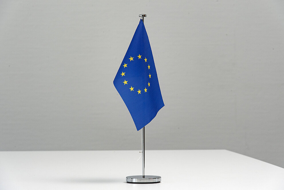 Auf einem weißen Tisch steht eine blaue EU-Flagge mit zwölf goldenen Sternen. Diese sind in einem Kreis angeordnet.