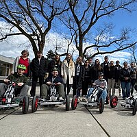 Eine Gruppe von Menschen steht im Freien und blickt in die Kamera; davor sitzen vier Kinder in kleinen Elektro-Karts.