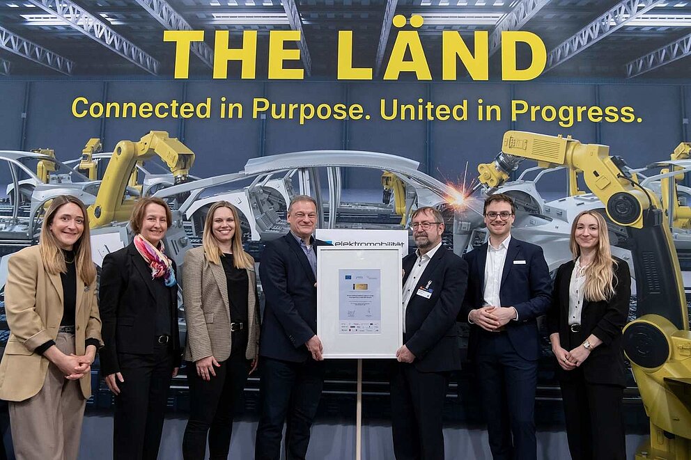 Sieben Personen stehen auf einer Messe vor einem Aufsteller, darunter Mitarbeitende des Clusters Elektromobilität Süd-West sowie der Geschäftsführer der e-mobil BW, Franz Loogen. In der Mitte wird die Auszeichnung ‚Gold Label‘ an das Cluster übergeben.