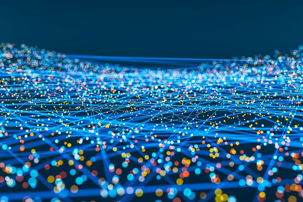 Abstrakte Darstellung eines digitalen Netzwerks: Viele bunte Lichtpunkte sind über ein Gitter aus leuchtend blauen Linien miteinander verbunden, vor dunklem Hintergrund.