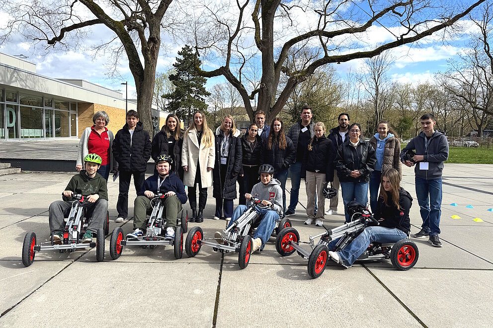 Eine Gruppe von Menschen steht im Freien und blickt in die Kamera; davor sitzen vier Kinder in kleinen Elektro-Karts.
