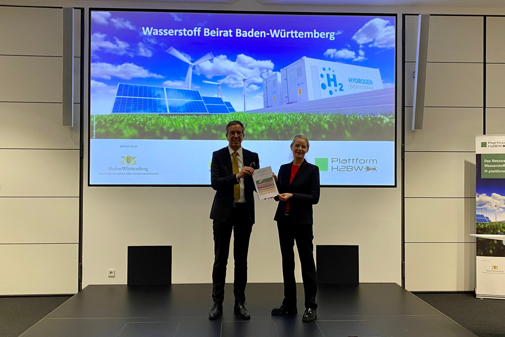 Prof. Markus Hölzle übergibt Umweltministerin Walker ein Printpapier auf der Bühne. Sie lächeln dabei ins Publikum.