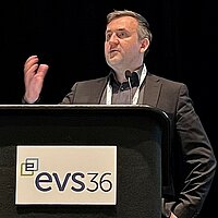 Dr. Wolfgang Fischer steht an einem schwarzen Rednerpult mit der Aufschrift "EVS36", er hebt erklärend die Hand 