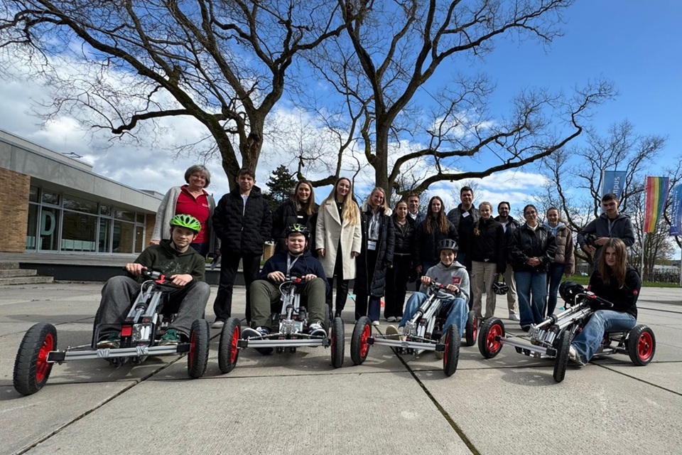 Eine Gruppe von Menschen steht im Freien und blickt in die Kamera; davor sitzen vier Kinder in kleinen Elektro-Karts.