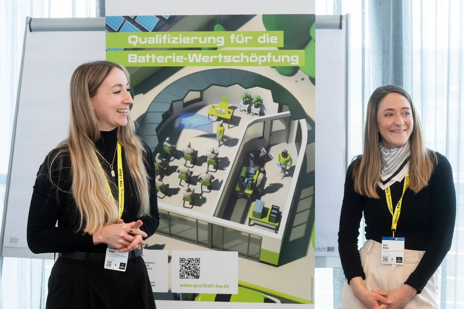 Zwei lächelnde Frauen stehen vor einem Roll-up mit der Aufschrift 'Qualifizierung für die Batterie-Wertschöpfung' und einer schematischen Darstellung eines Batterie-Ökosystems.